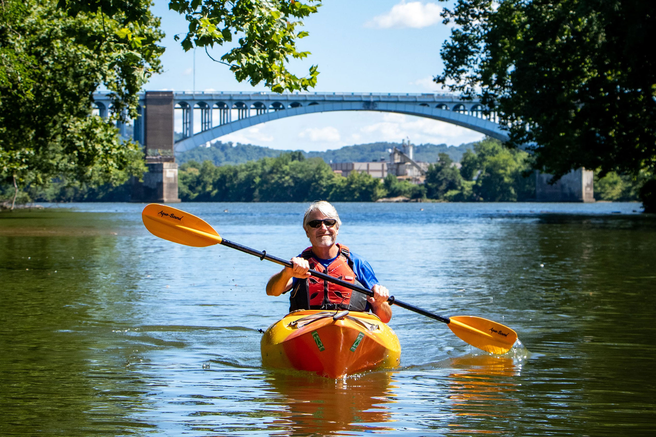 Smiling man in orange kayak paddles down a river in Pittsburgh.